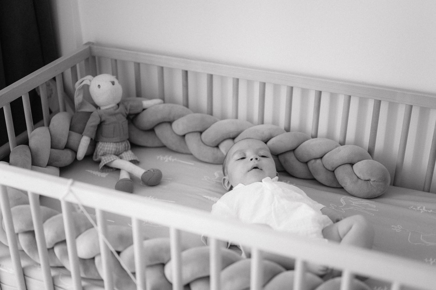 Black and white photograph showing a crib with a stuffed monkey toy and braided bumper padding.