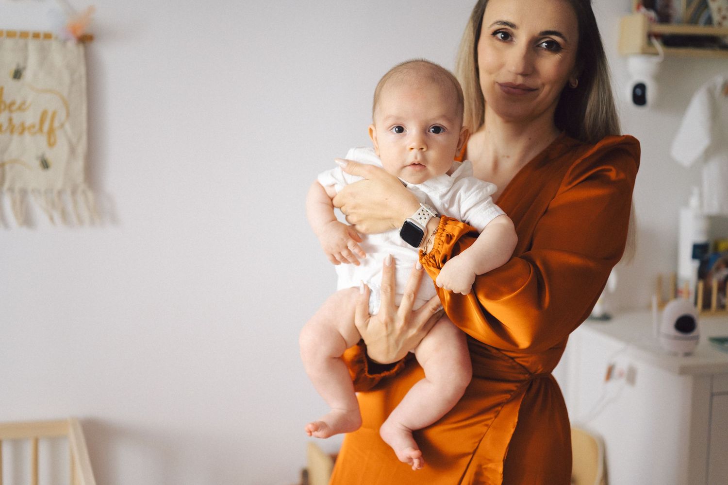 Someone holds a baby wearing white clothes while standing in a cozy room decorated with neutral colored furnishings.