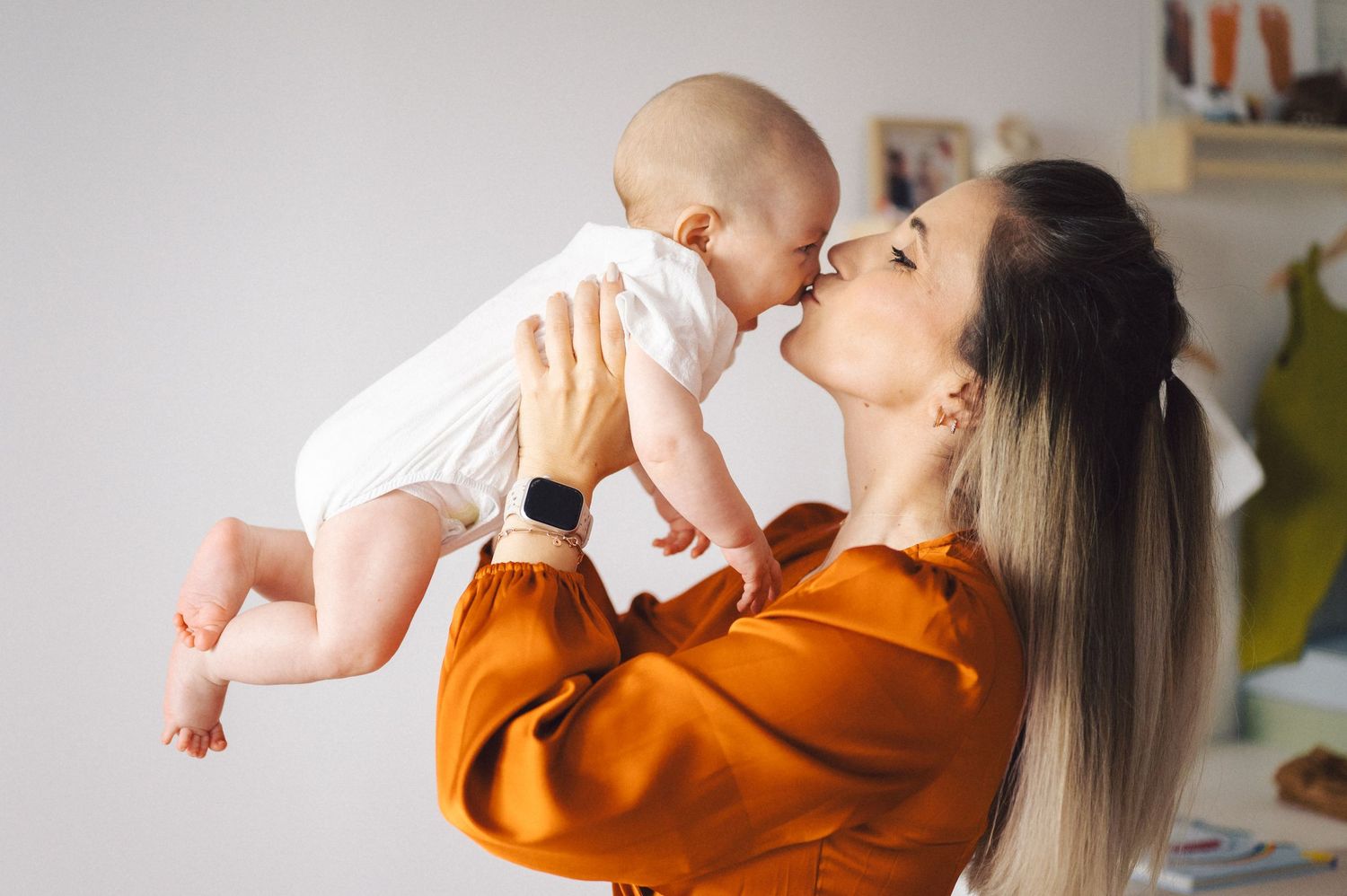 A joyful moment is captured as a parent lifts and kisses a baby wearing a rust-colored outfit against a light interior.
