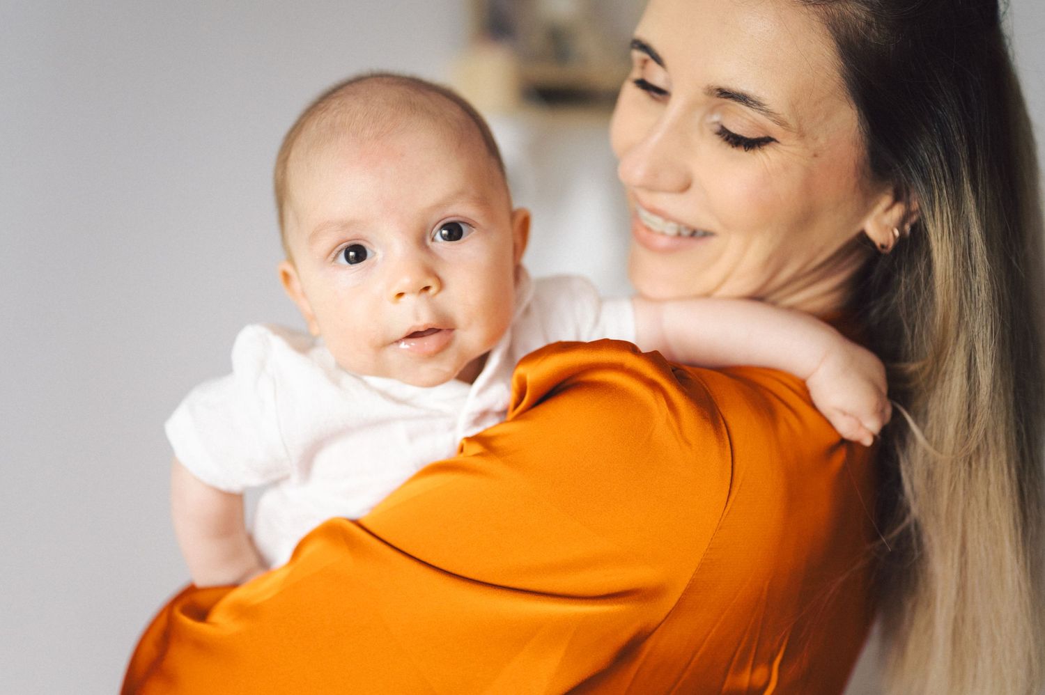 A baby in a white outfit rests against someone wearing a bright orange sweater in a tender moment.