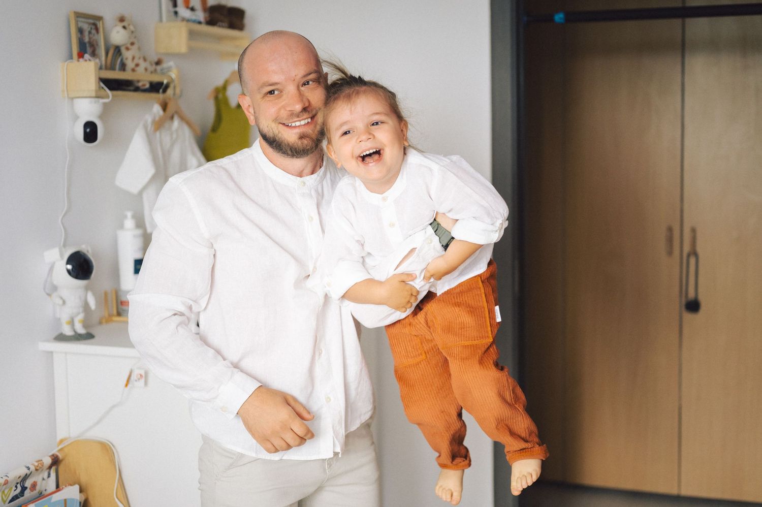 Parent in white shirt holds a giggling child wearing an orange outfit in a bright indoor setting.