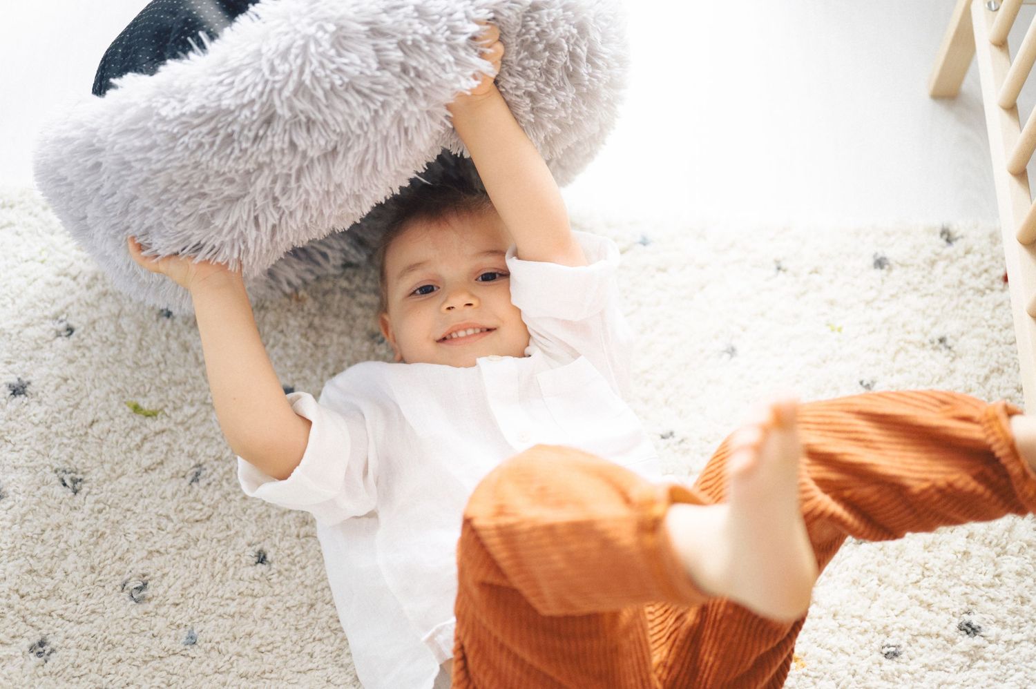 A small figure holds up a fluffy gray pillow while lying on a white carpet, wearing orange pants and a white shirt.