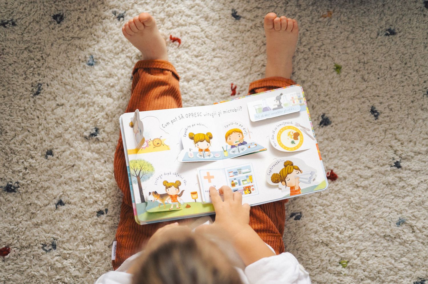 Baby's feet and hands visible while lying on carpet reading a colorful children's board book.