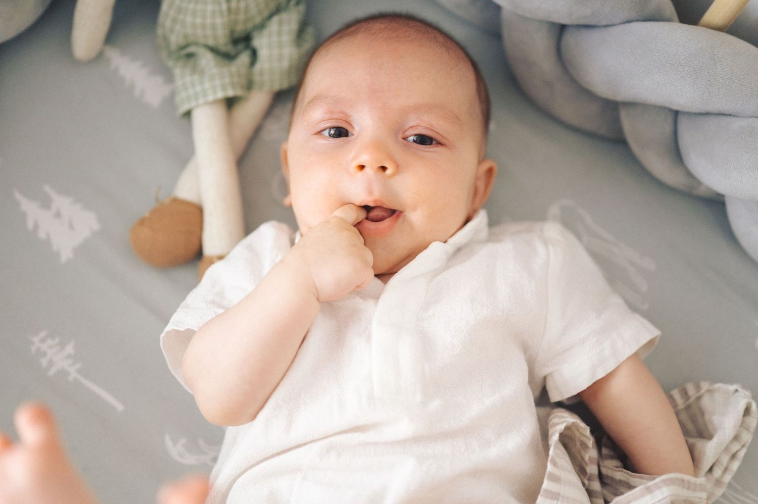 A sweet baby in a white onesie lies on a soft blanket surrounded by plush toys.