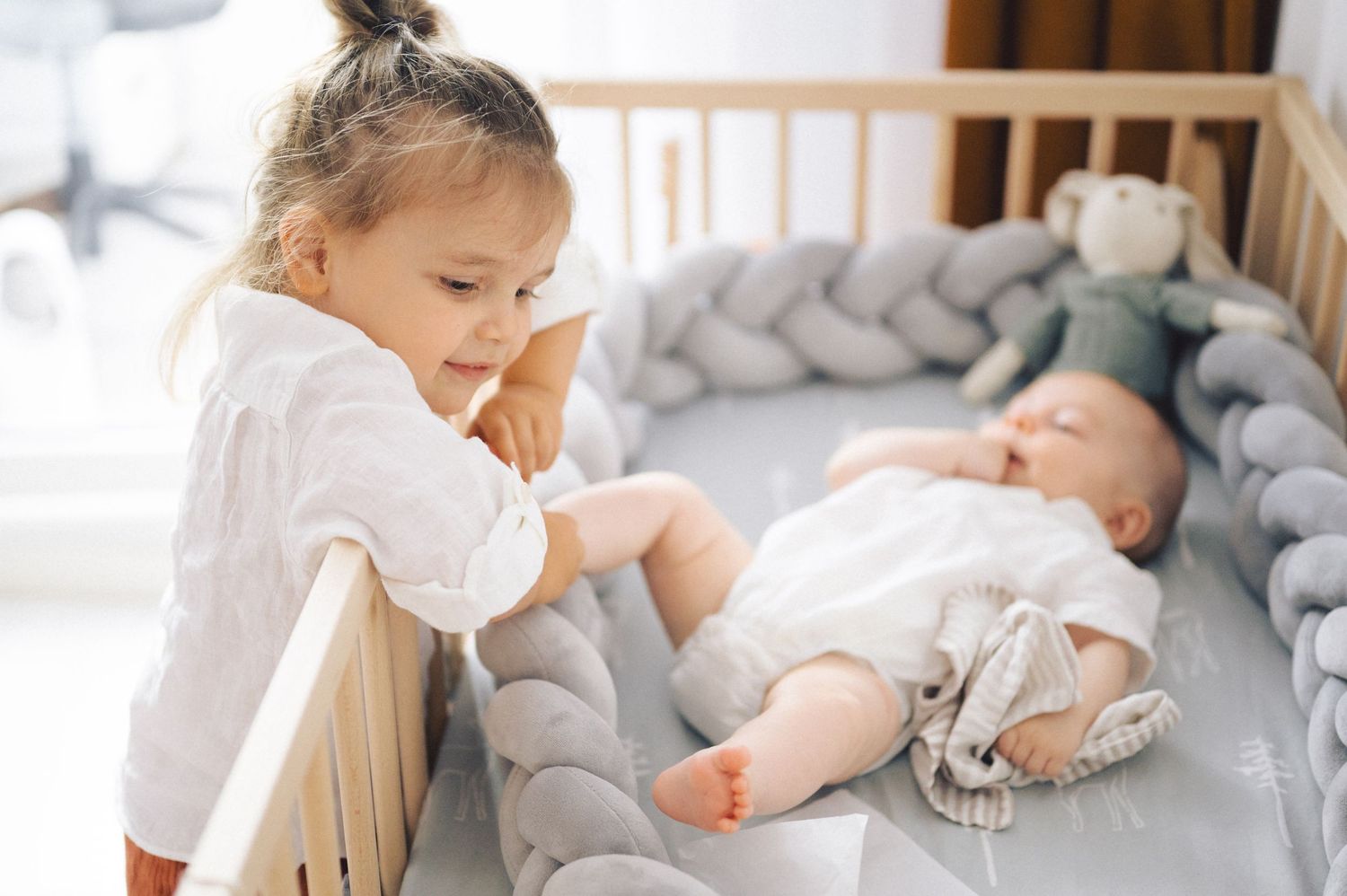 Two young children lie on white bedding while playing with a gray cat creating a heartwarming home scene.