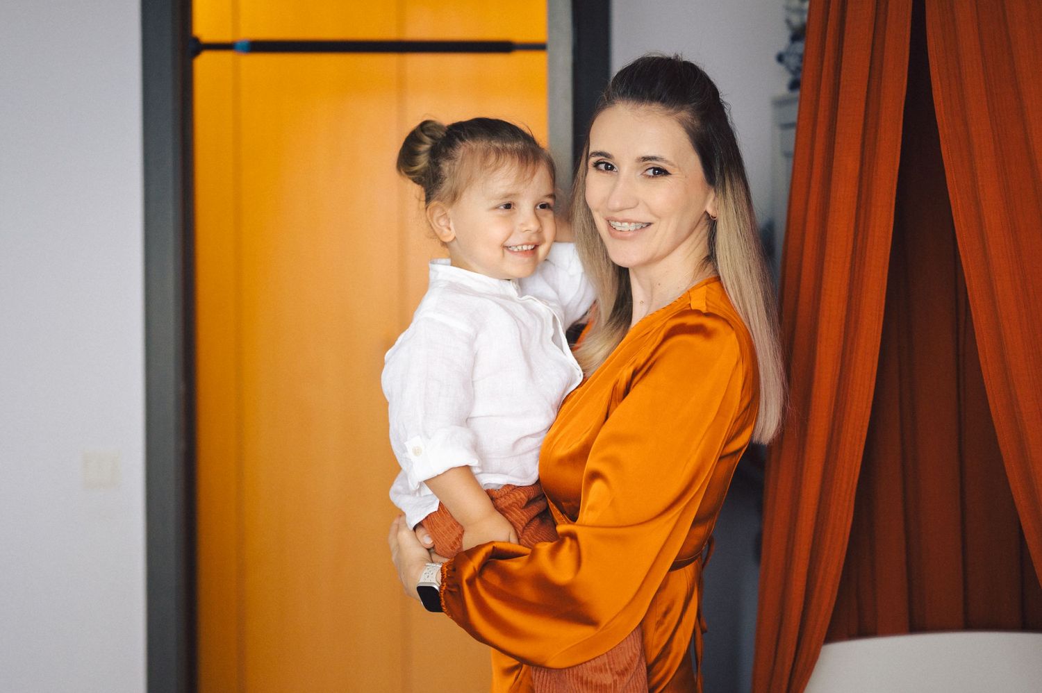 Adult in bright orange blazer holding small child wearing white top against orange and grey background.
