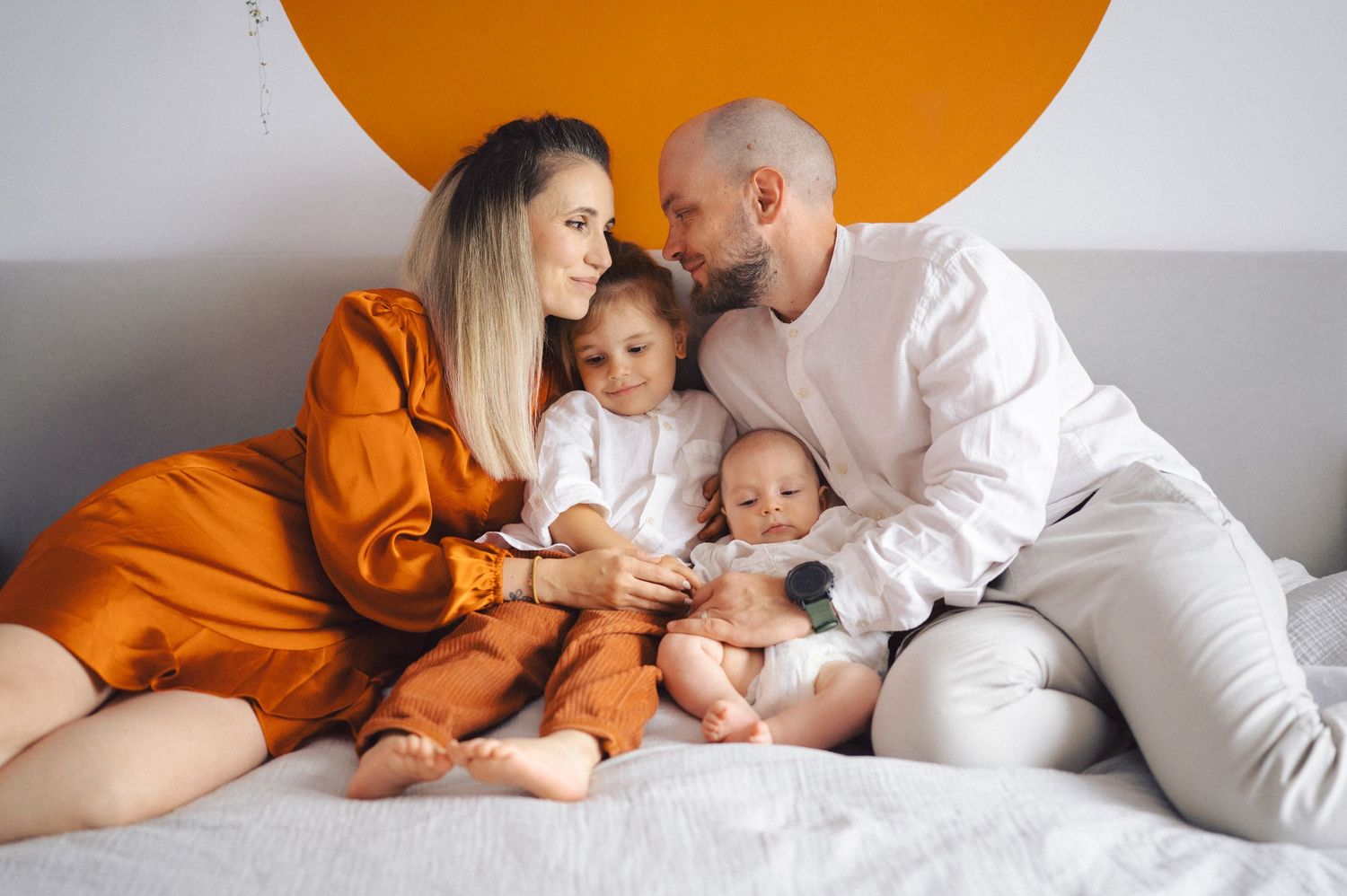 A family group lounges together on a white couch beneath a large orange circular wall decoration.