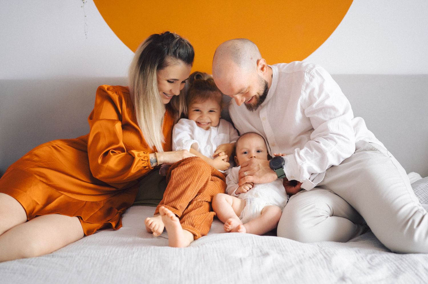 Family lying together on light backdrop in orange and white outfits in sweet candid moments during studio photo session.