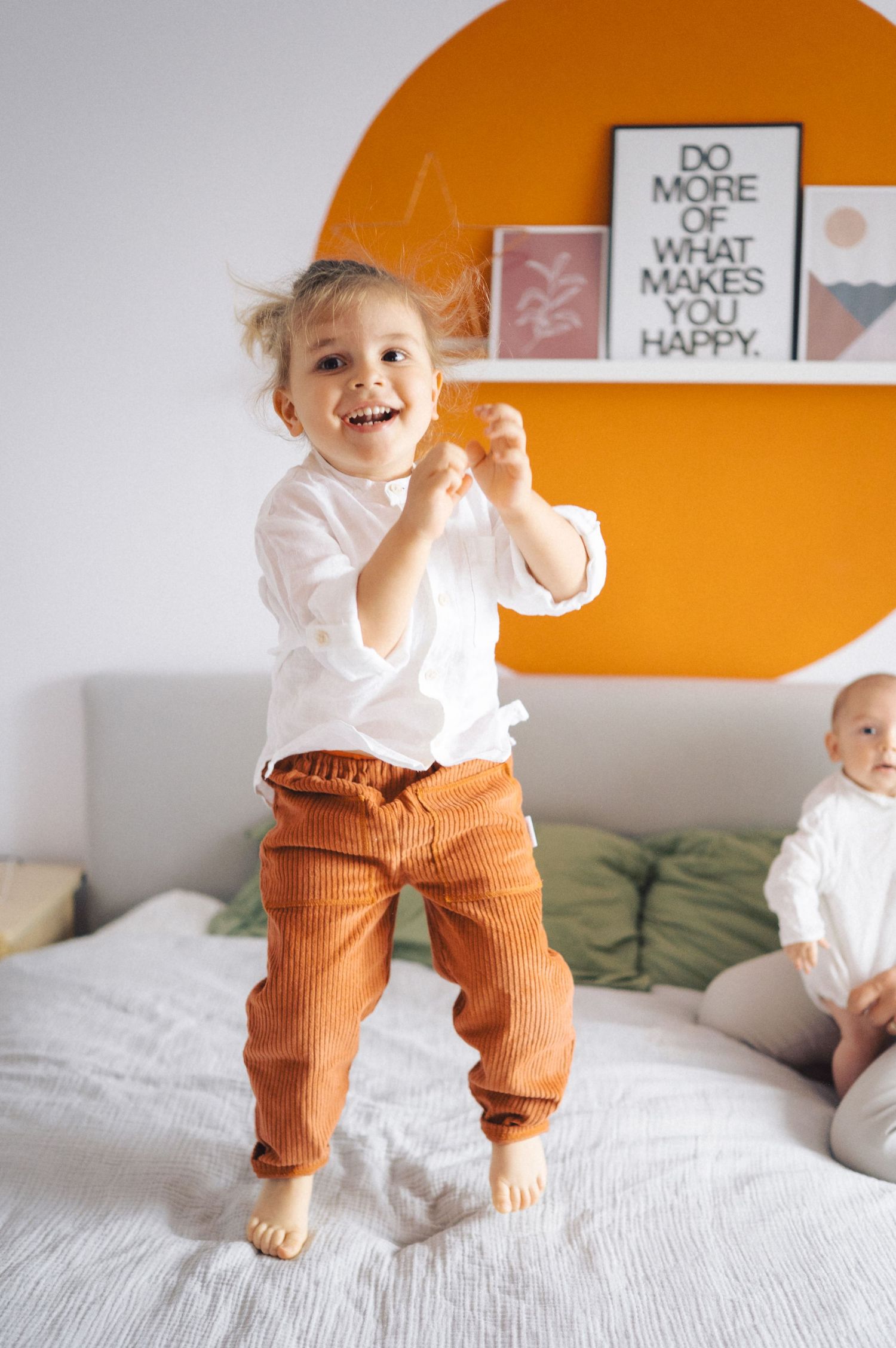 A joyful child jumps on a white bed wearing orange pants and white shirt against an orange circle wall accent.