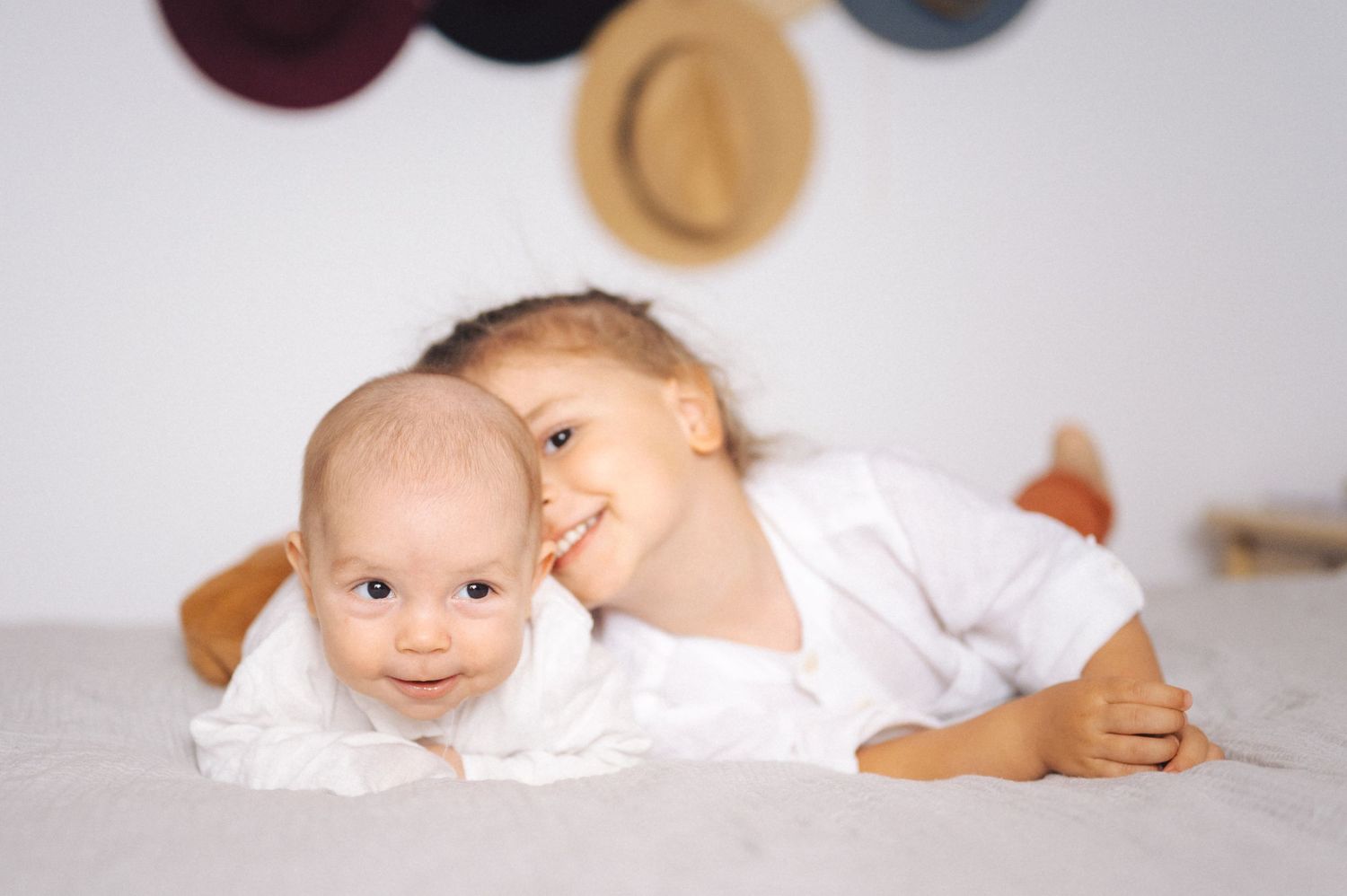 Two babies in white clothing lying together on a white blanket with decorative hats hanging on the wall.