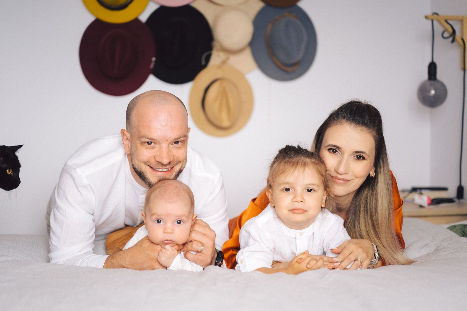A family lies together on a white surface with decorative hats displayed on the wall behind them.