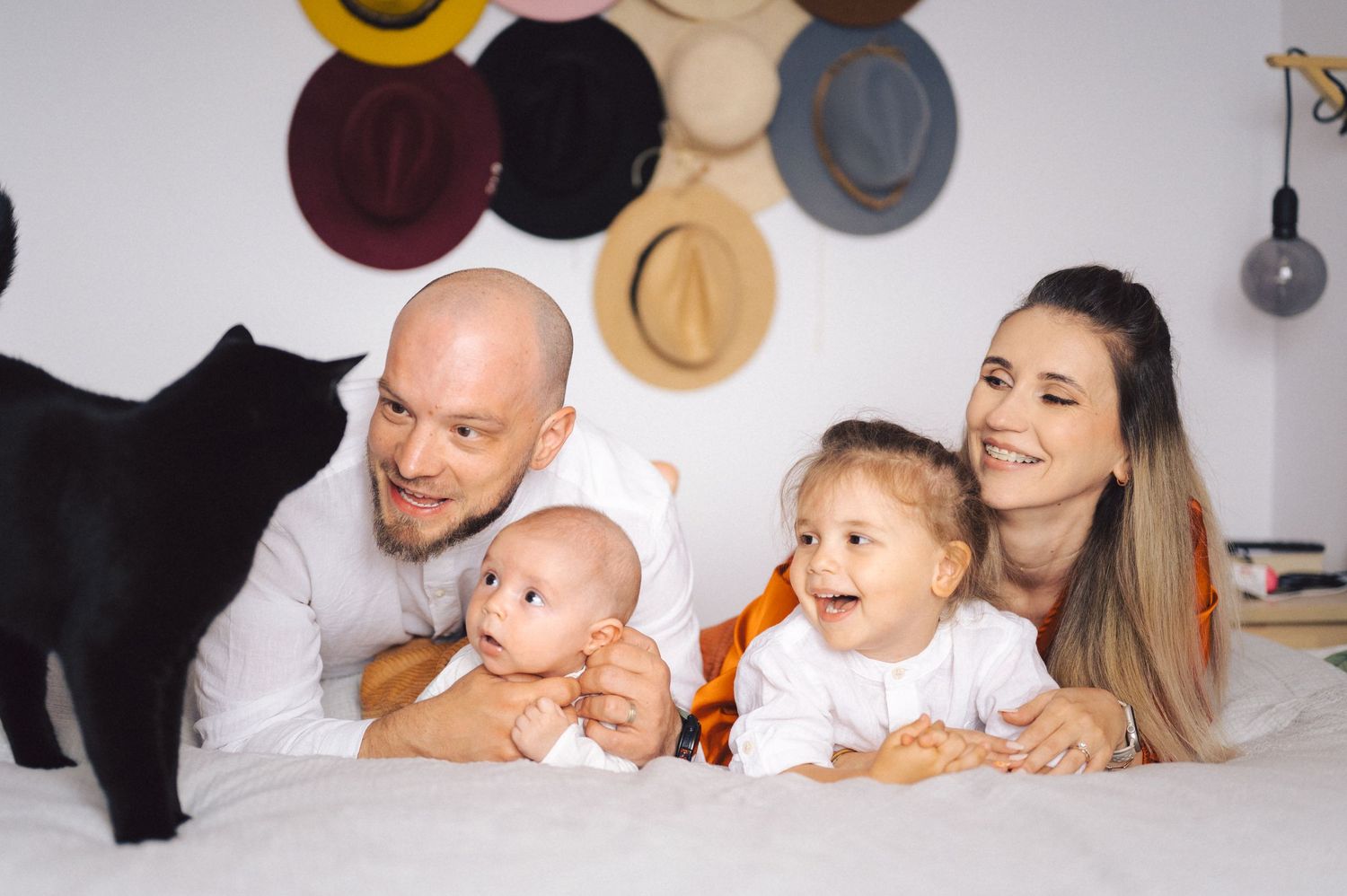 A family lies together on a white bed while hats hang decoratively on the wall behind them.