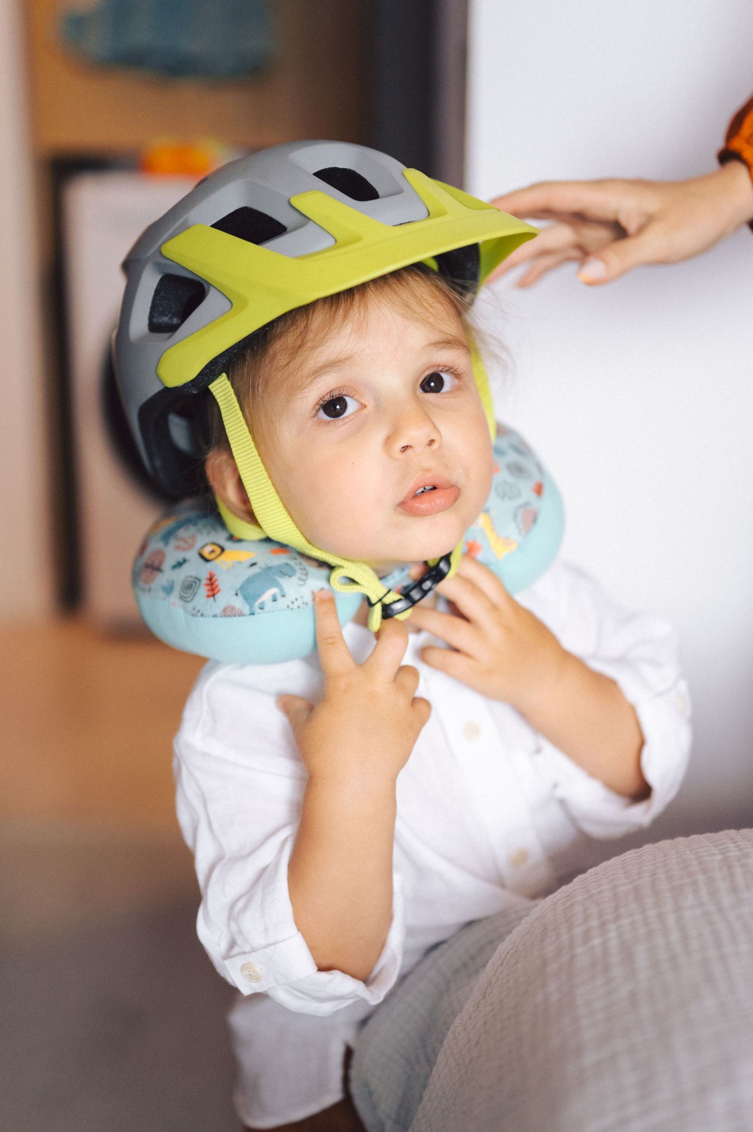 Child wearing yellow and gray bicycle helmet and white shirt sitting with an inflatable neck pillow.