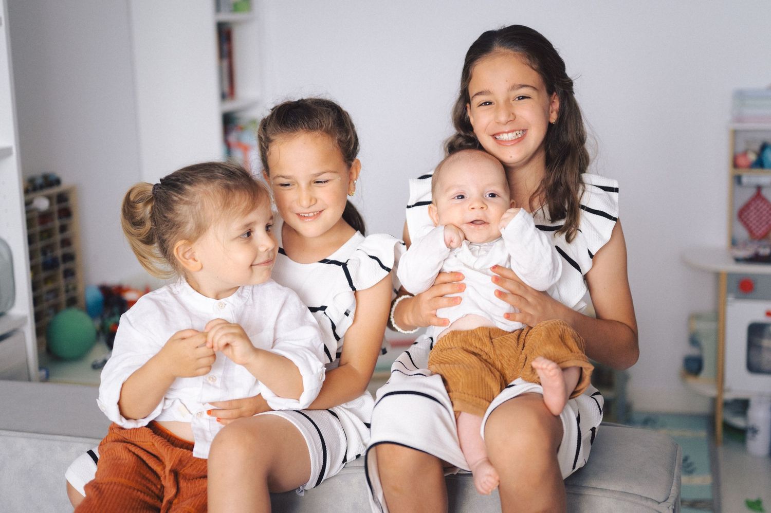 Four children in matching white and brown outfits sit together smiling on a couch in a bright room.