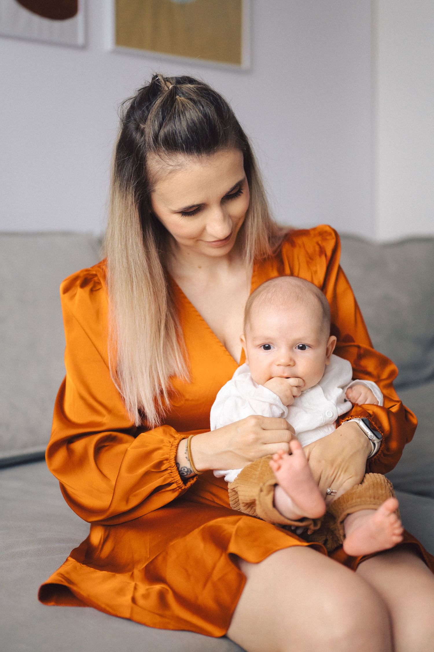 Mother in orange silk robe tenderly holds infant baby while sitting on couch in minimalist home setting.