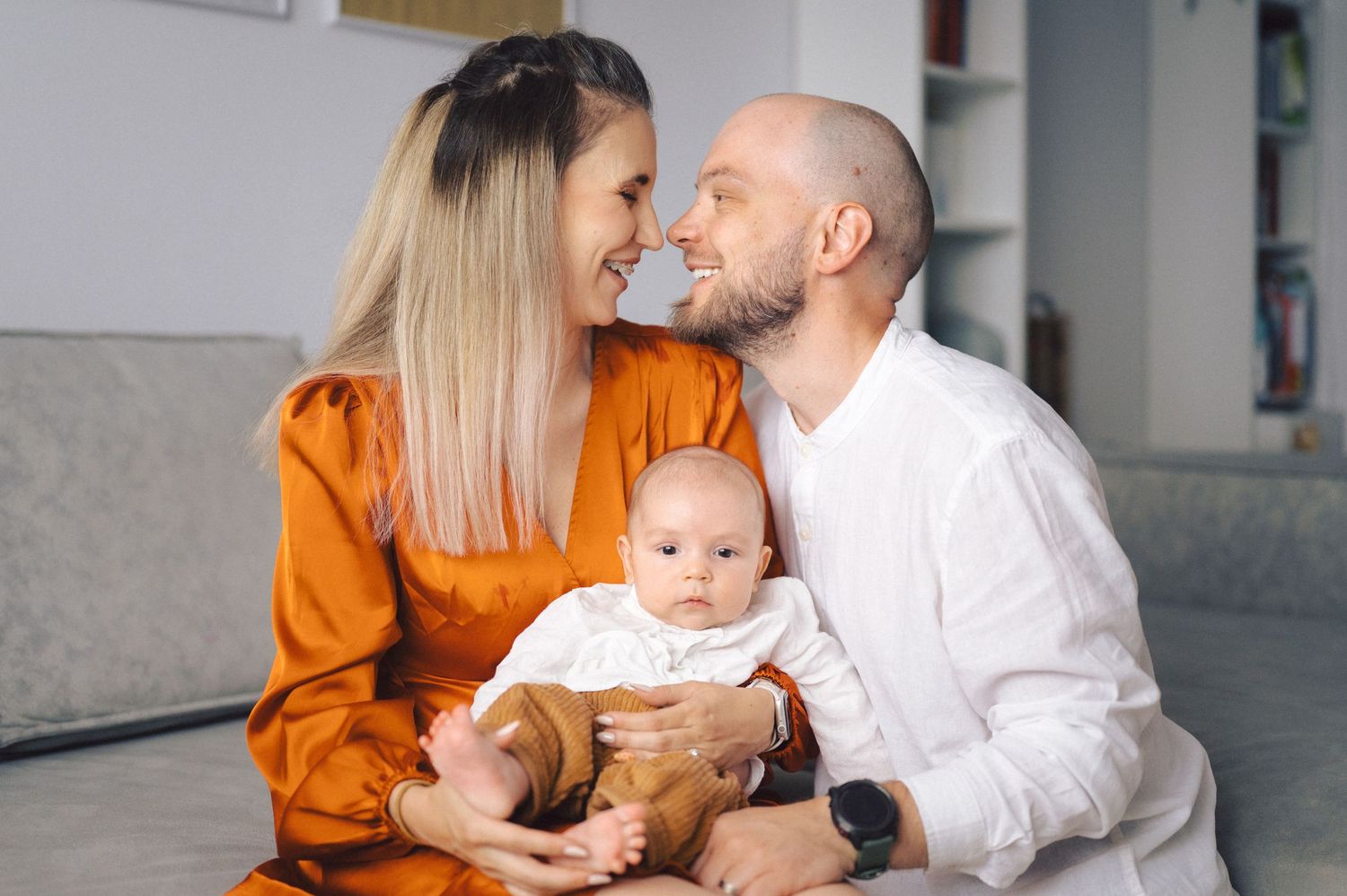 Couple sharing tender moment while holding baby in orange and white outfits at home.