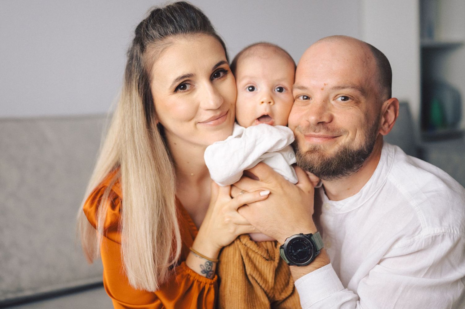 Parents showing affection while holding a baby and posing for tender family portraits in a bright indoor setting.