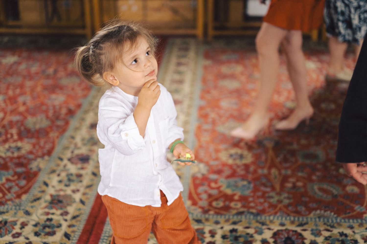 A small child in white shirt and orange pants stands thoughtfully on a patterned rug.