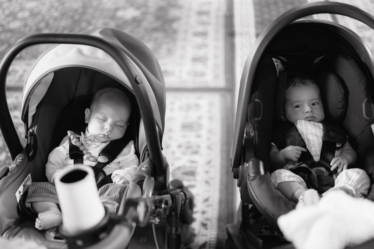 Two infants sit in identical car seats, with one holding what appears to be a coffee mug in this black and white photograph.