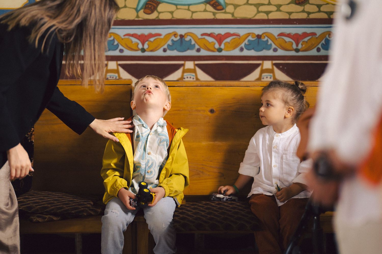 Two children sit on a bench with decorative wall designs behind them while someone gestures nearby.