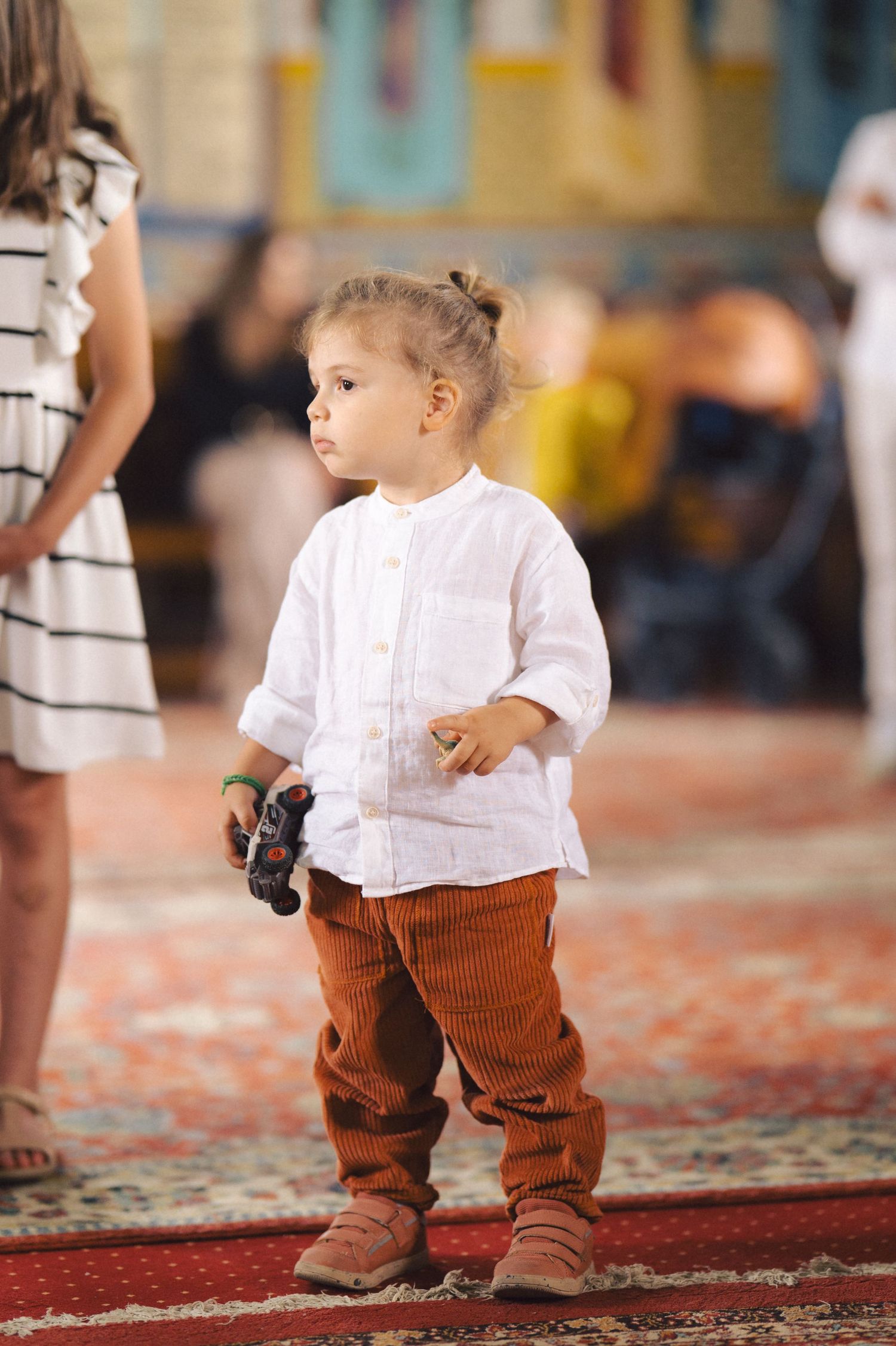 Young toddler in white shirt and brown corduroy pants stands on patterned carpet while holding a toy.