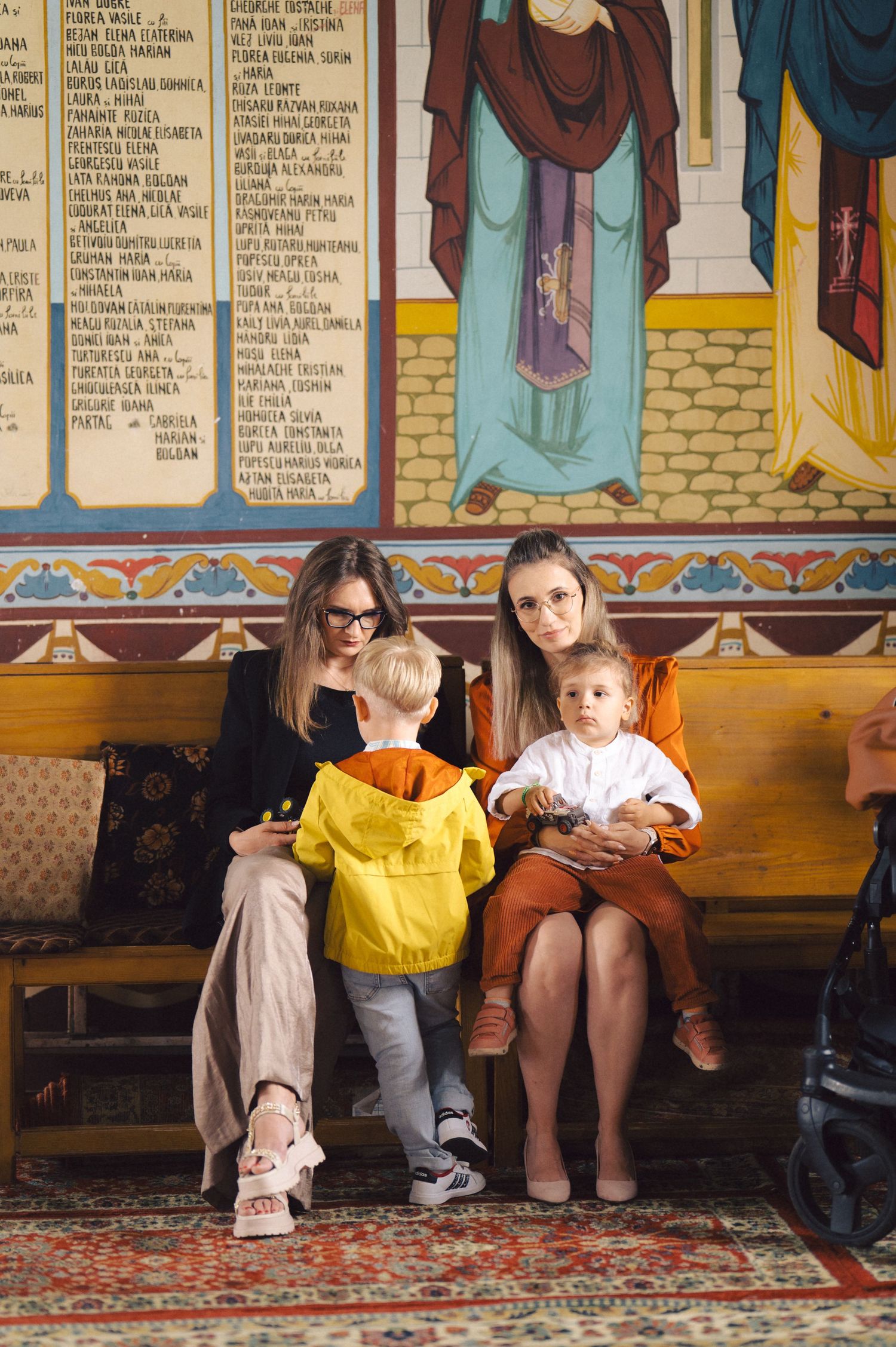 Adults and children sit together on a bench beneath decorative wall art in an ornate interior setting.