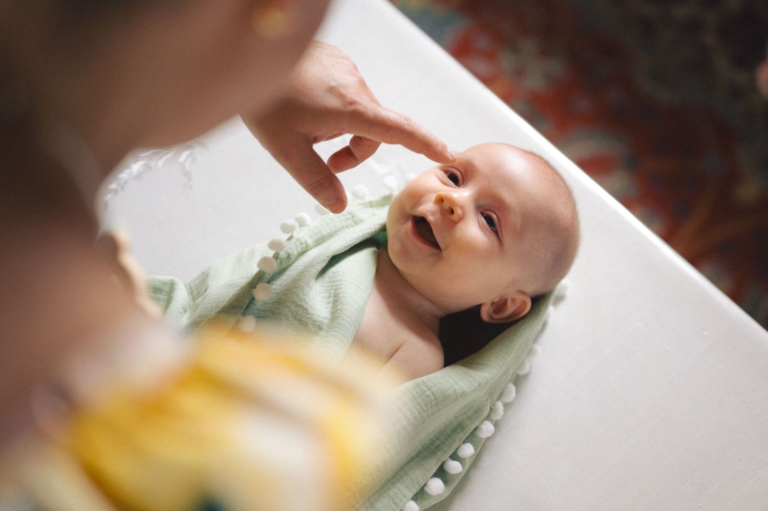 A happy smiling baby lying down during a diaper change with a caregiver's hand visible in the frame.