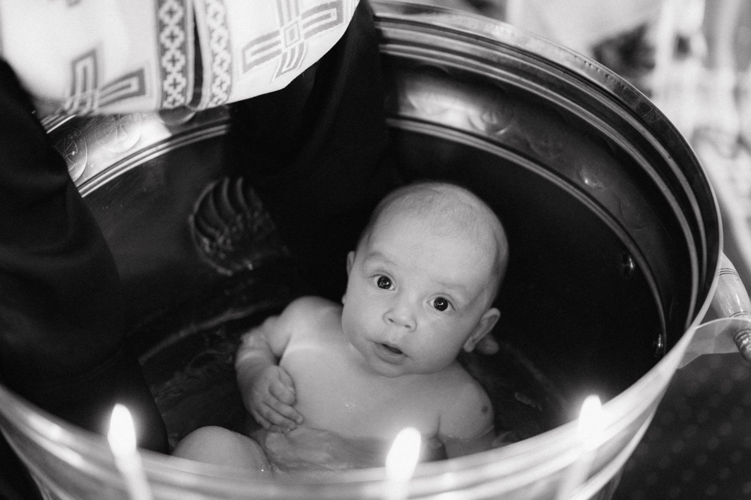 A black and white photo shows a small baby sitting in a vintage pram or carriage looking up at the camera.