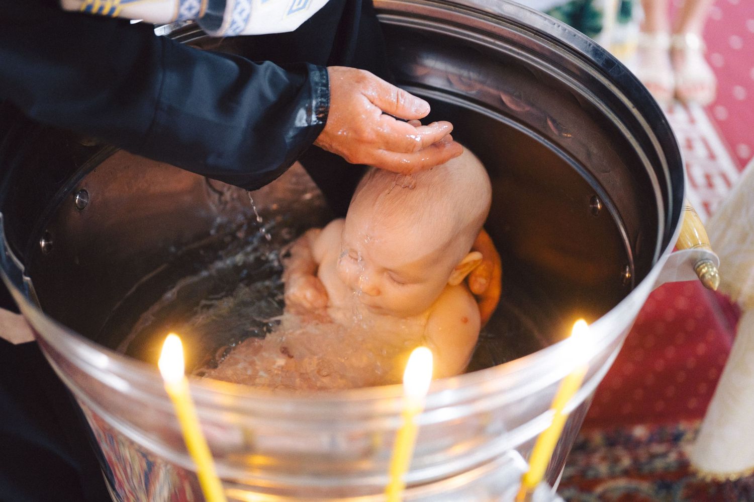 A hand pouring water during a baptismal ceremony with lit candles around the baptismal font.