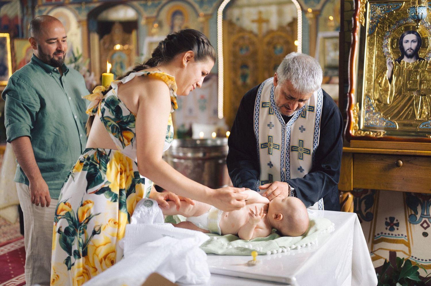 An infant baptism ceremony takes place in an ornate Orthodox church with a priest and family members present.