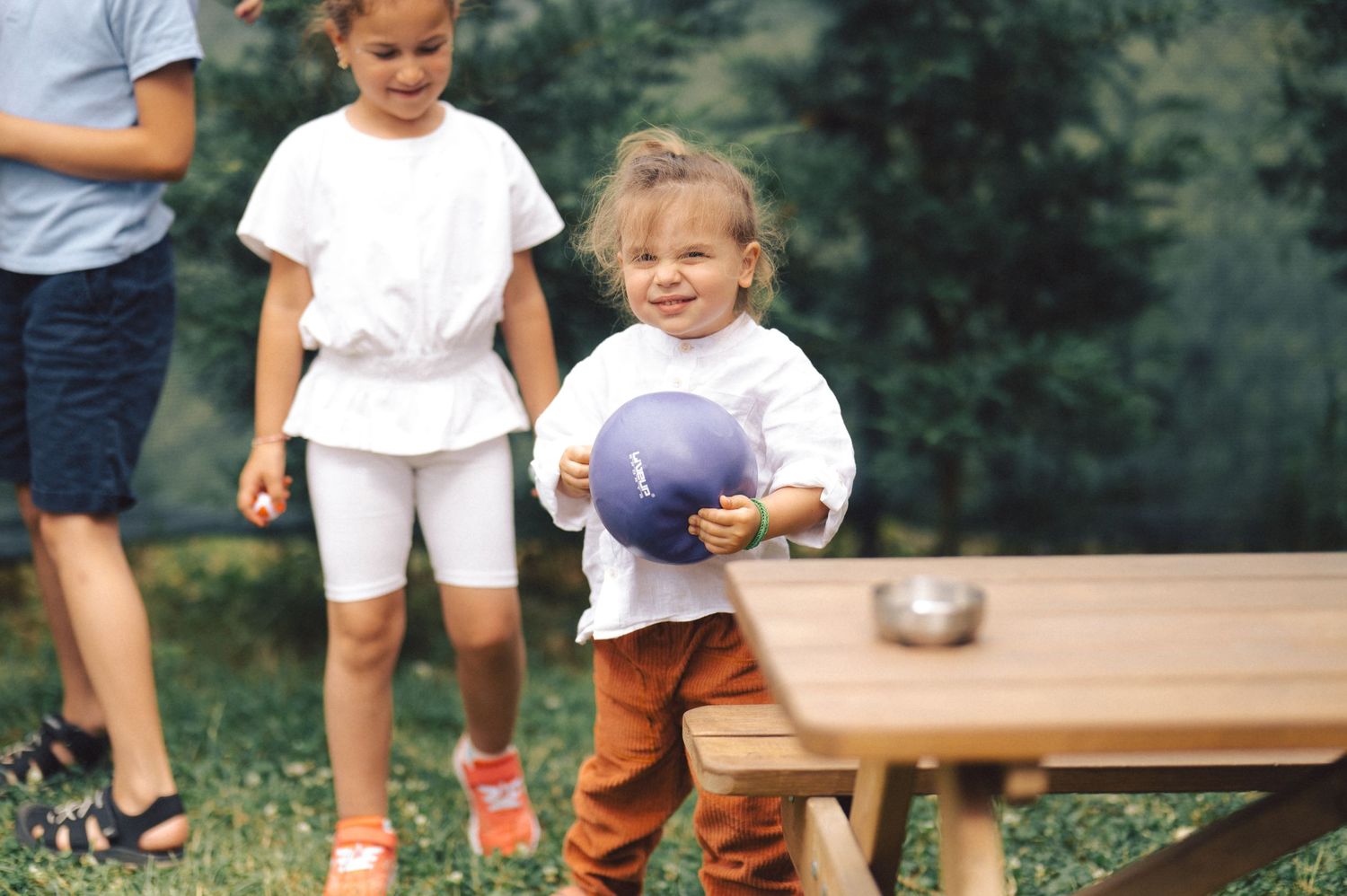 Children playing outdoors near a wooden picnic table with one holding a purple ball in casual summer attire.