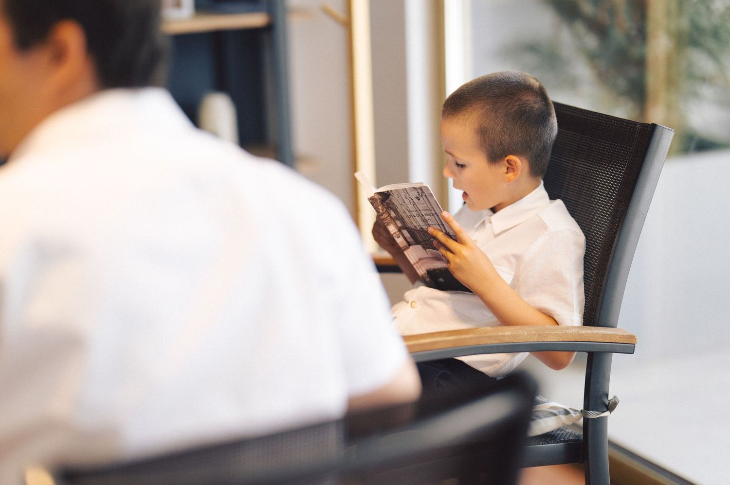 Young child in white shirt sits at desk intently focused on digital tablet device while holding with both hands.