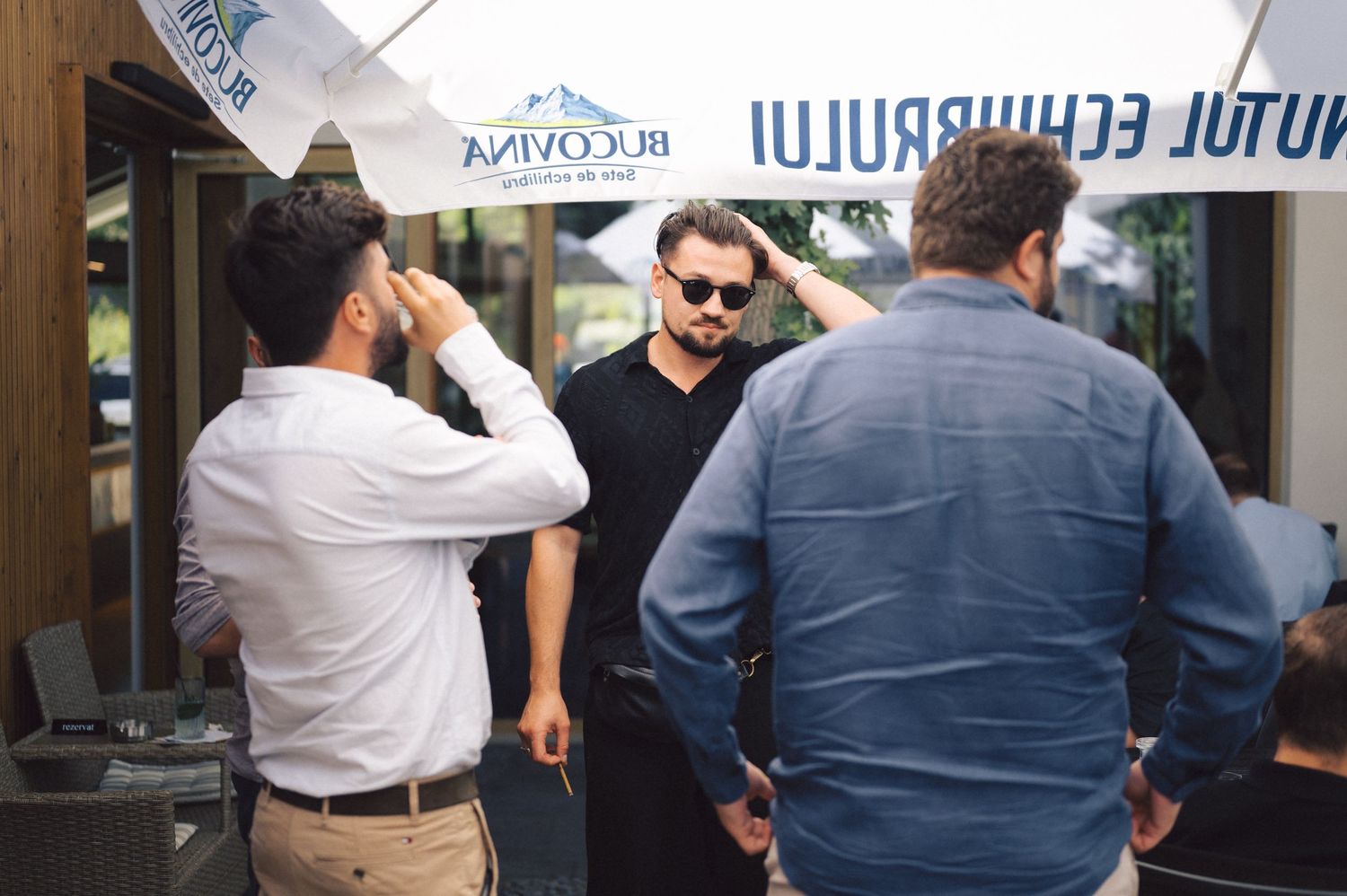 Three people stand and converse outside a restaurant or cafe under a white umbrella.