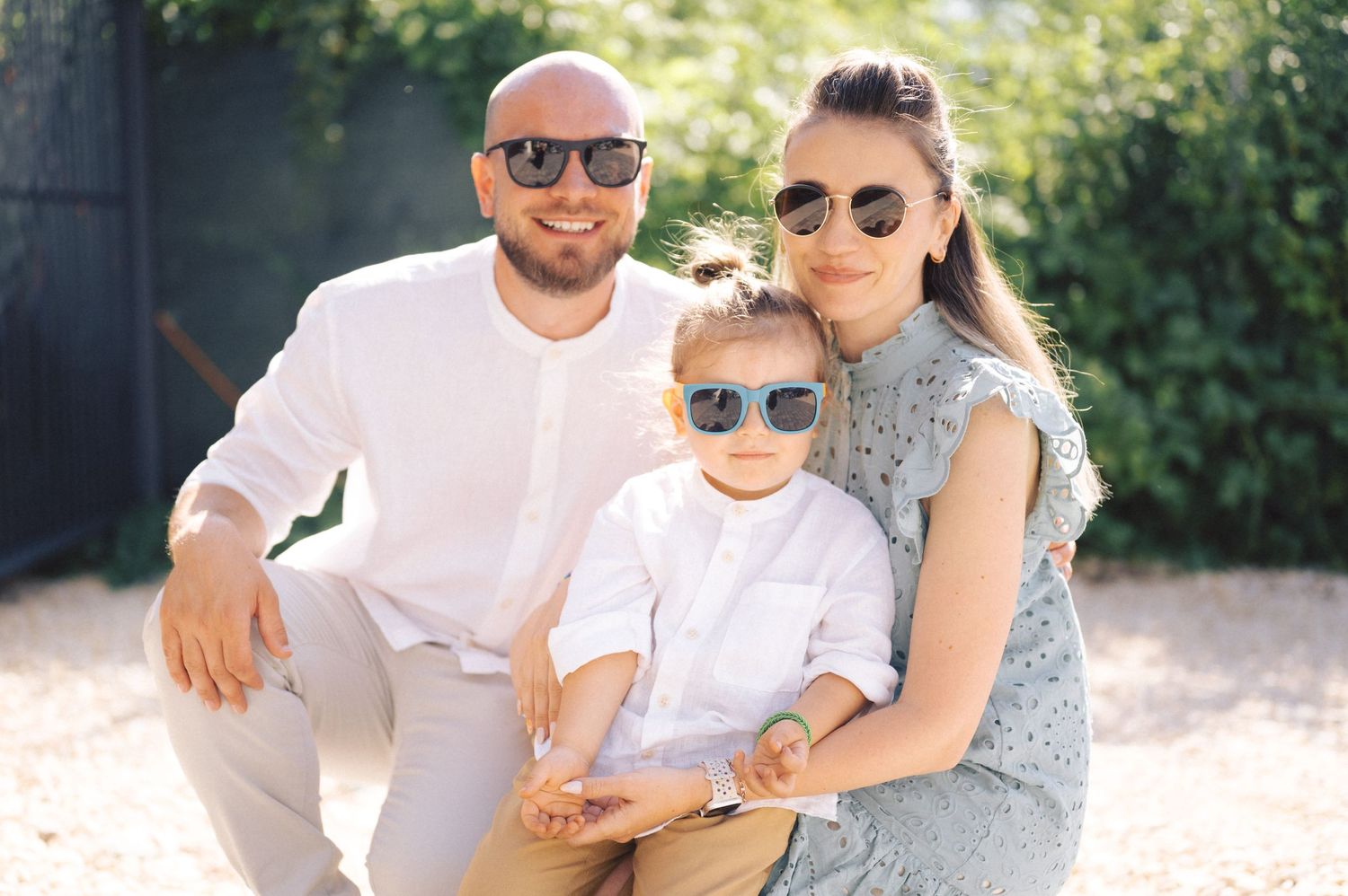 A family of three wearing sunglasses poses together outdoors in coordinated summer outfits.