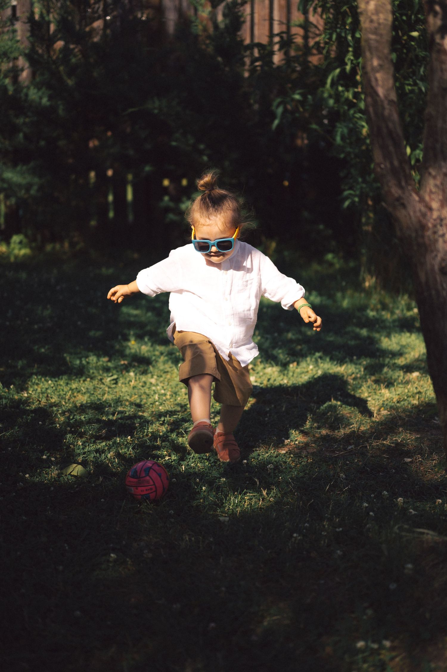A young child in a white dress explores shadows and sunlight while playing outdoors on a grassy hill.