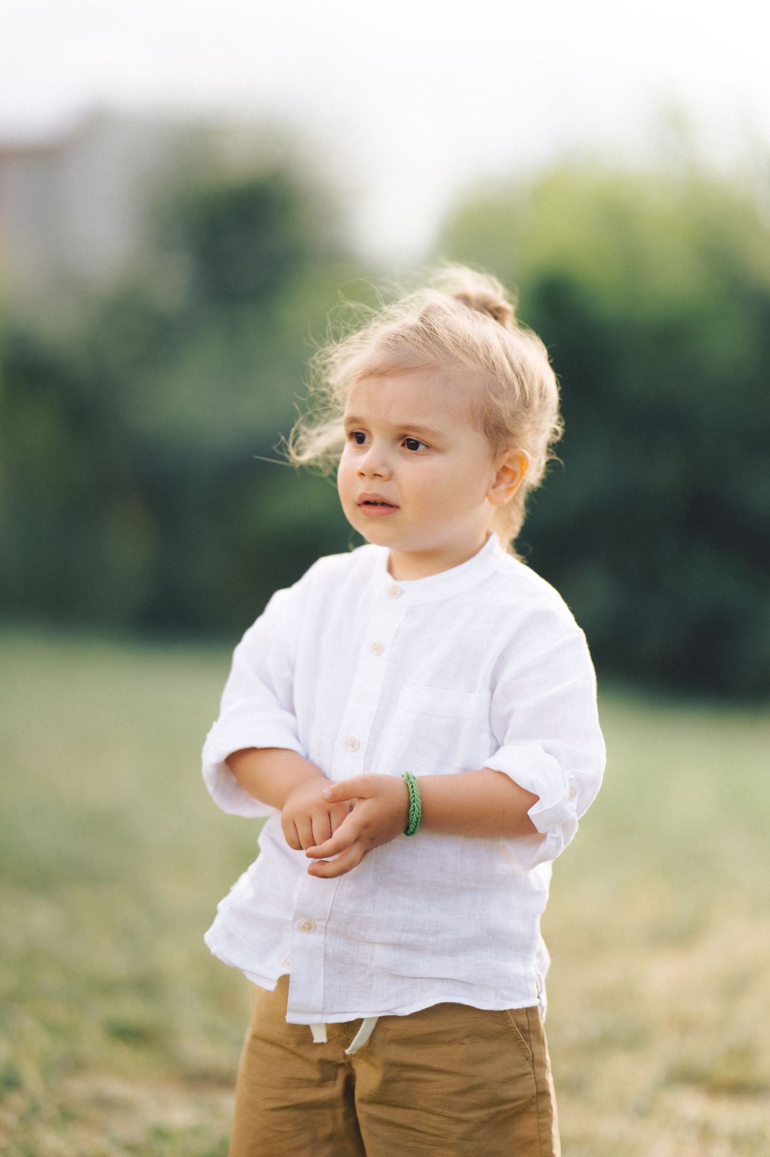 A young child in a white linen shirt and brown pants stands outdoors against a blurred nature background.
