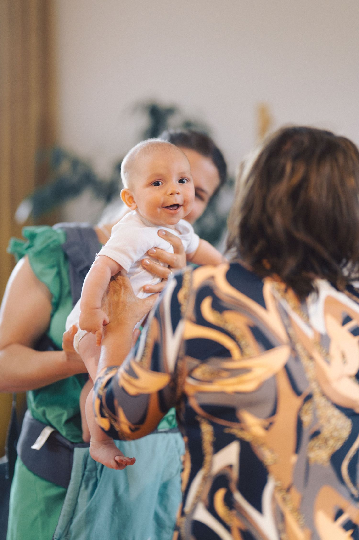 A smiling baby in a white onesie is held up while two people interact in an indoor setting.