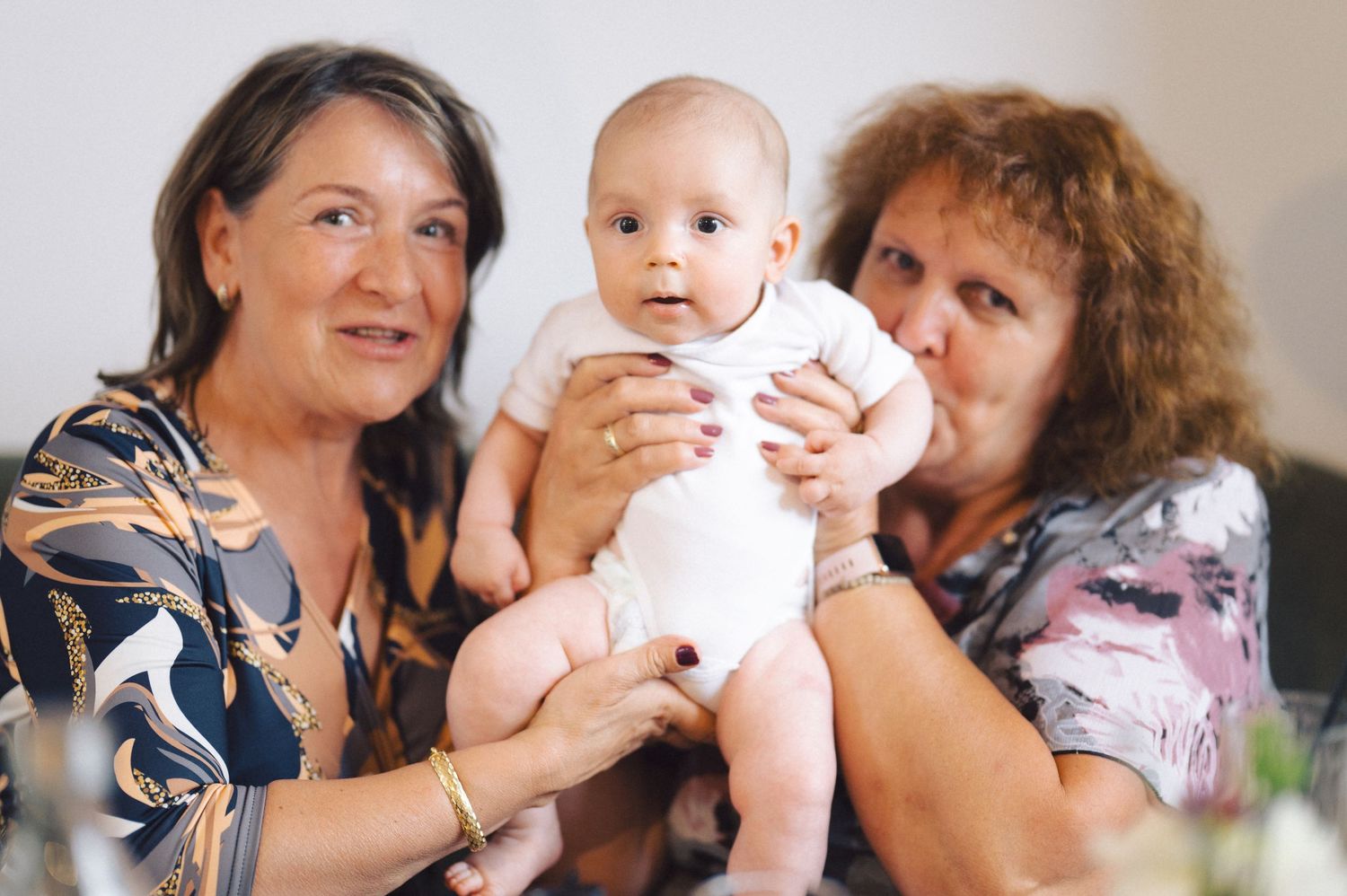 A baby in a white onesie is supported by two adults in a warm indoor setting.
