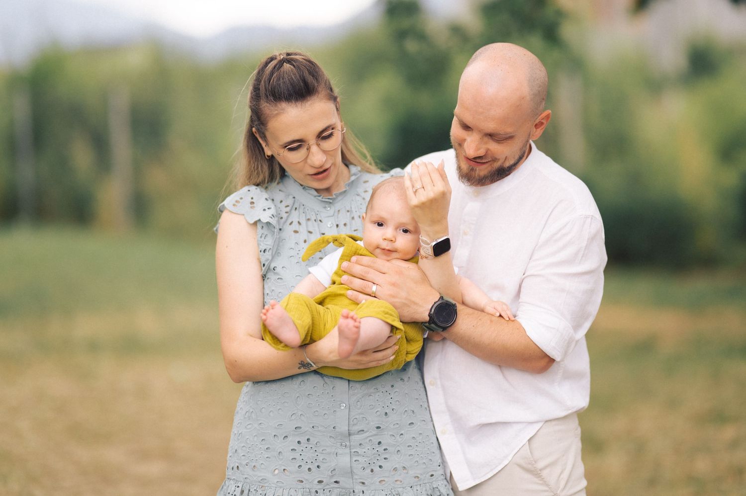 Family in grey dress and white shirt holding baby in yellow outfit in sunny outdoor field setting.