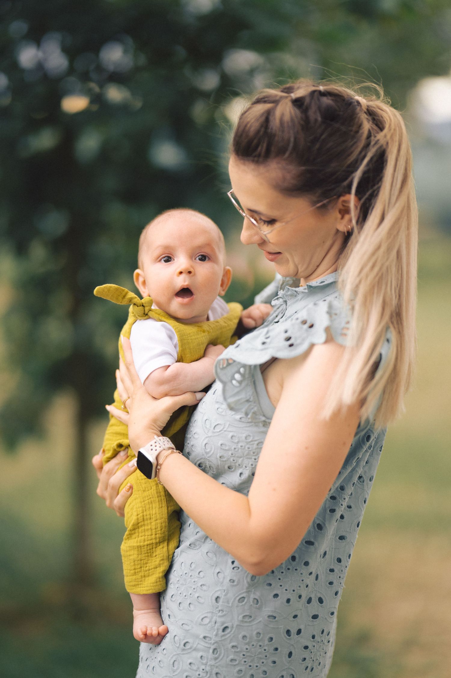 A loving interaction between a parent wearing a gray top and a baby in a yellow romper against a blurred outdoor background.