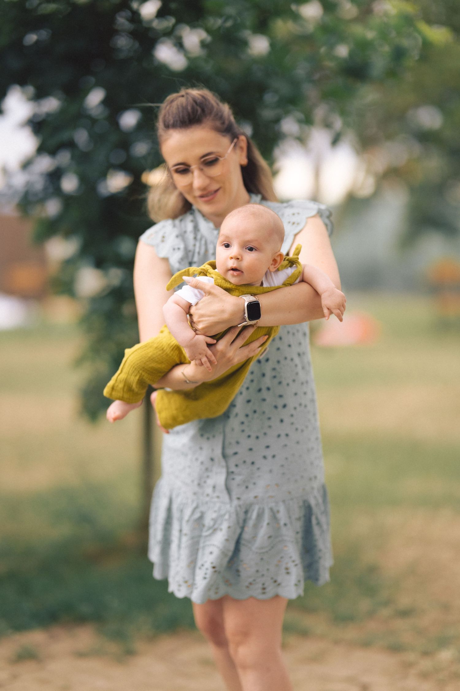 A parent in a gray dress holds and lovingly embraces a baby wearing a yellow outfit in an outdoor park setting.