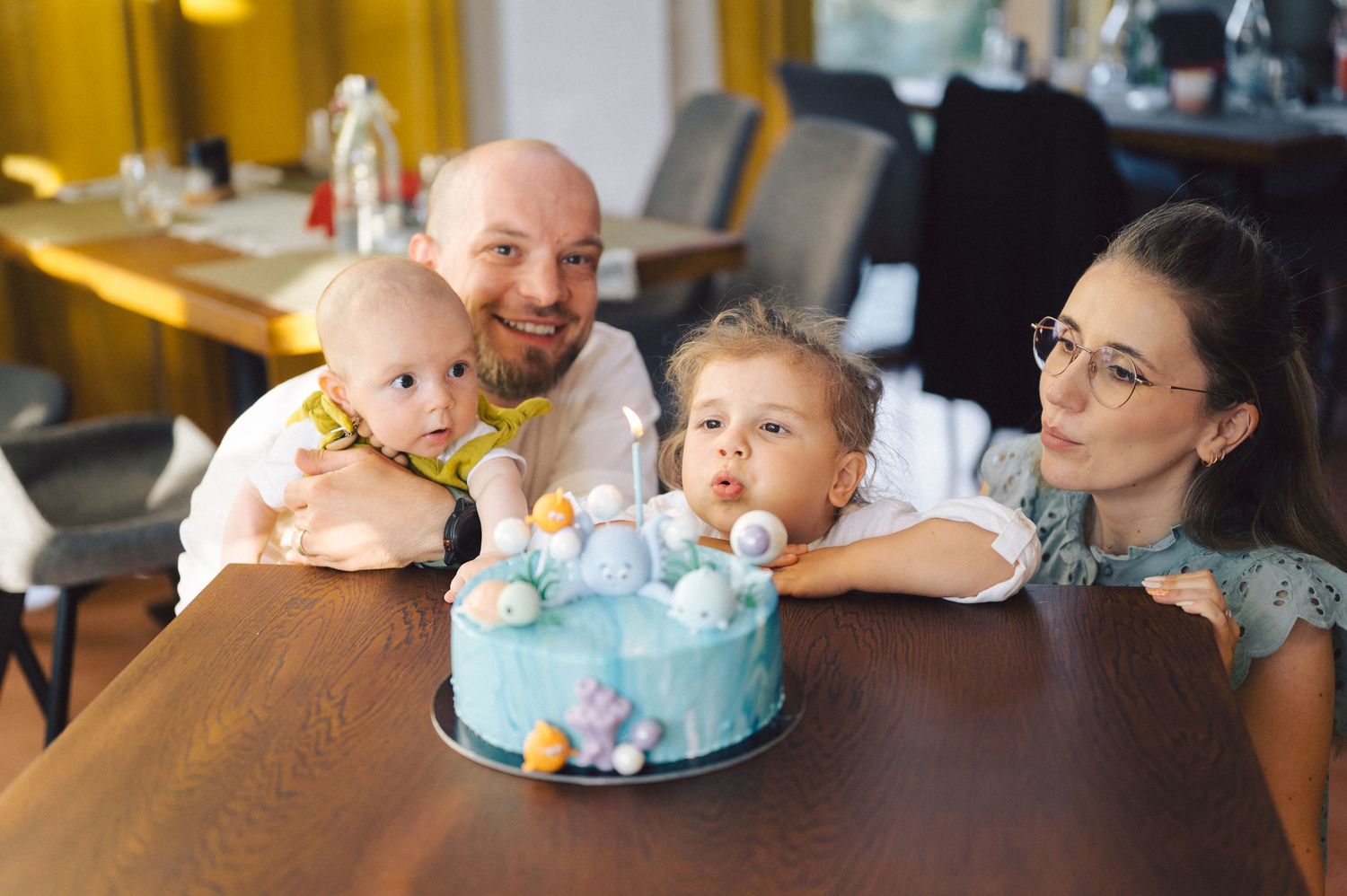 A family celebrates together around a blue birthday cake at a wooden table in a warm-lit dining area.
