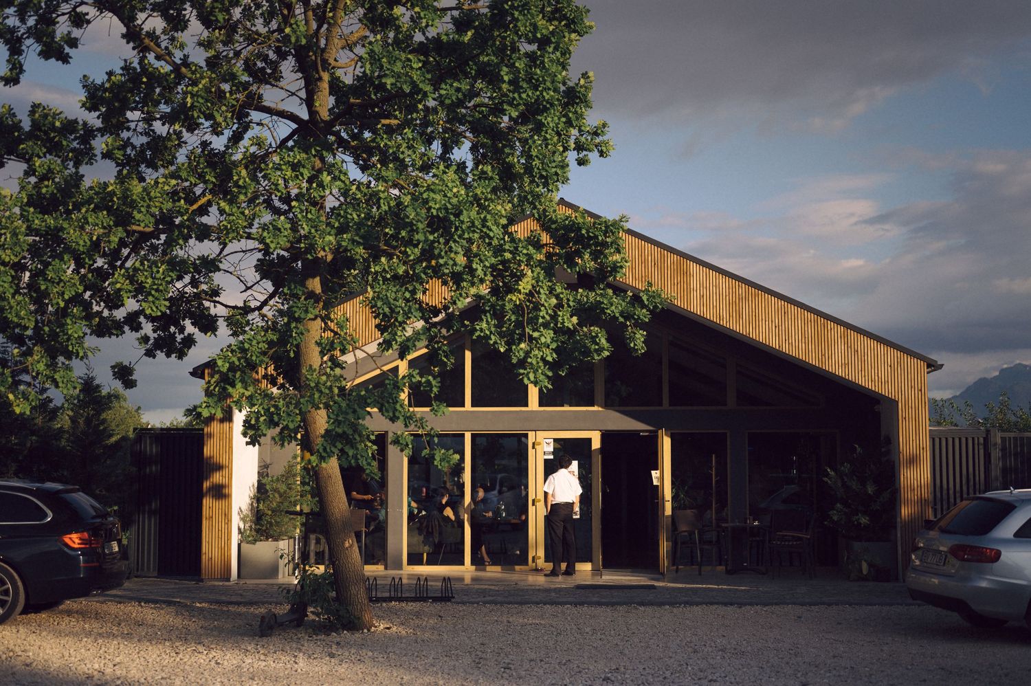 Modern wooden cafe building with large windows and outdoor seating area surrounded by parked cars and trees at sunset.