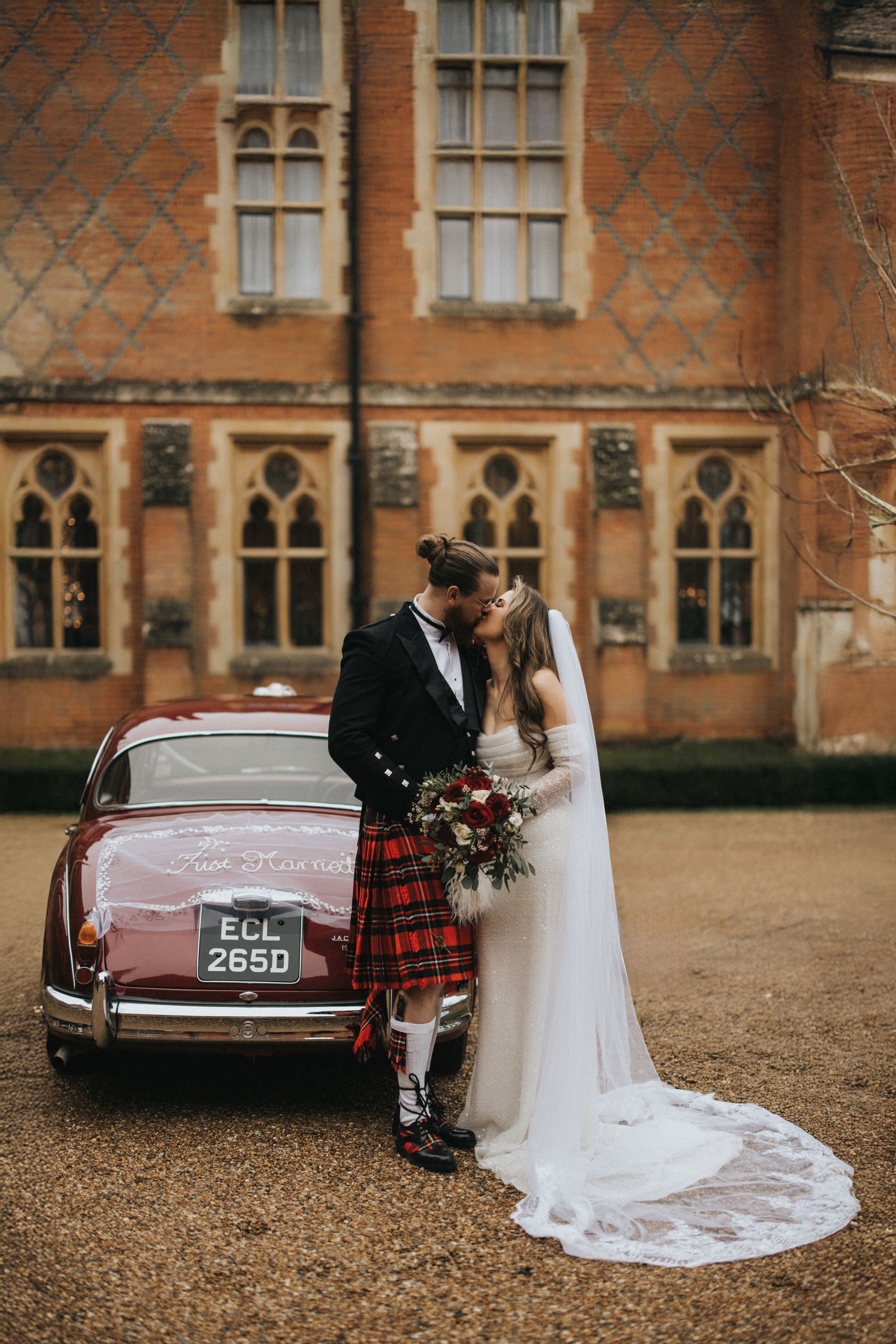 A couple in wedding attire stands by a vintage car outside a historic brick building with Gothic windows.