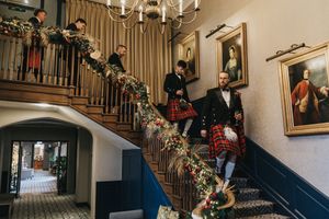 Members of a wedding party in traditional Scottish kilts descend an elegant staircase adorned with holiday garland.