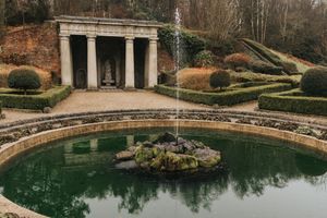 Classical garden pool with columned pavilion and ornate fountain surrounded by manicured hedges in a formal estate setting.