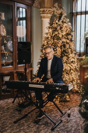 A musician performs on a digital keyboard in an elegant room with a beautifully lit Christmas tree in the background.