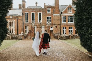 A couple walks along a gravel path outside a historic brick manor house, with one wearing a tartan kilt and white dress.
