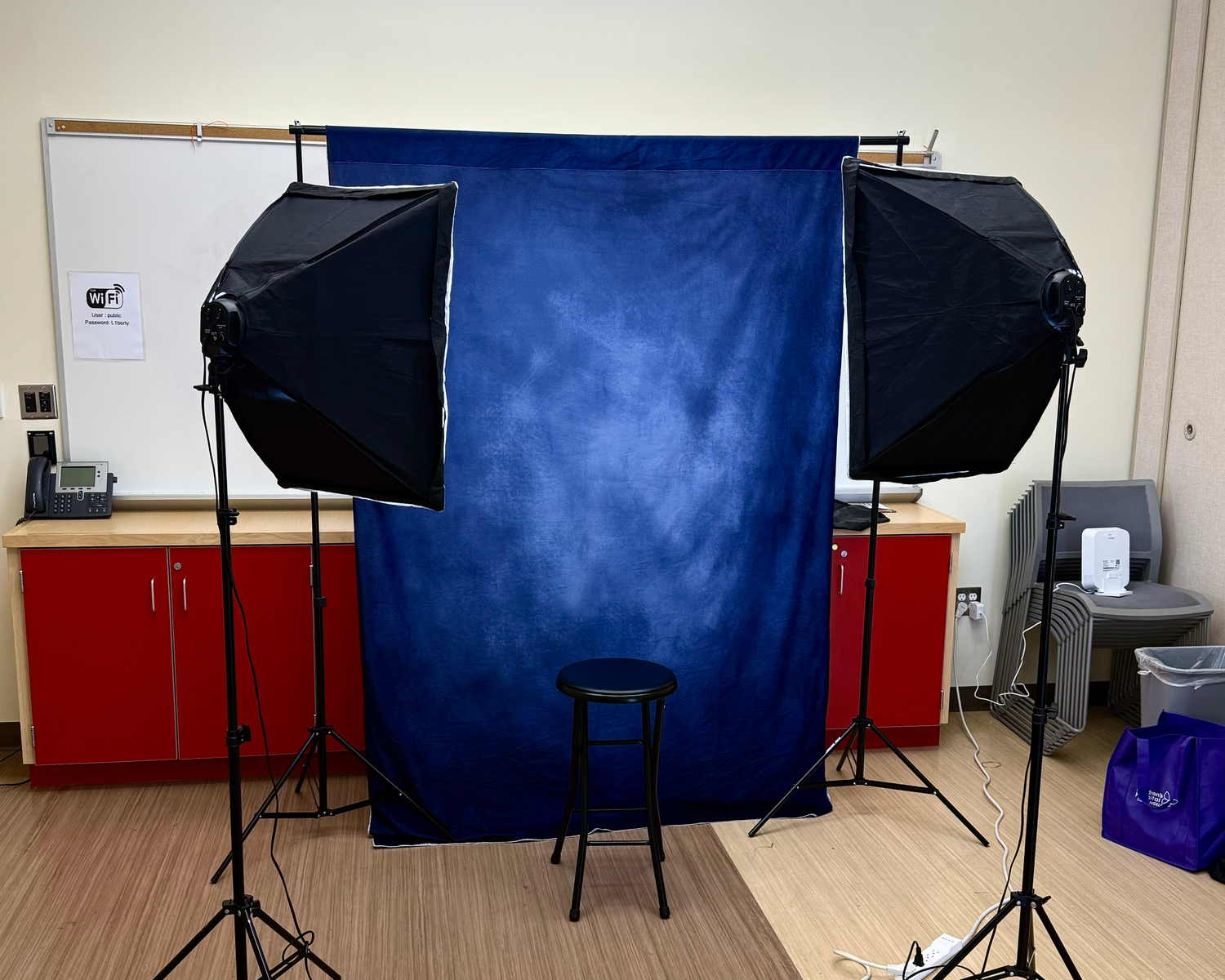 Photo studio setup with blue backdrop, stool, and two softbox lights in a classroom.