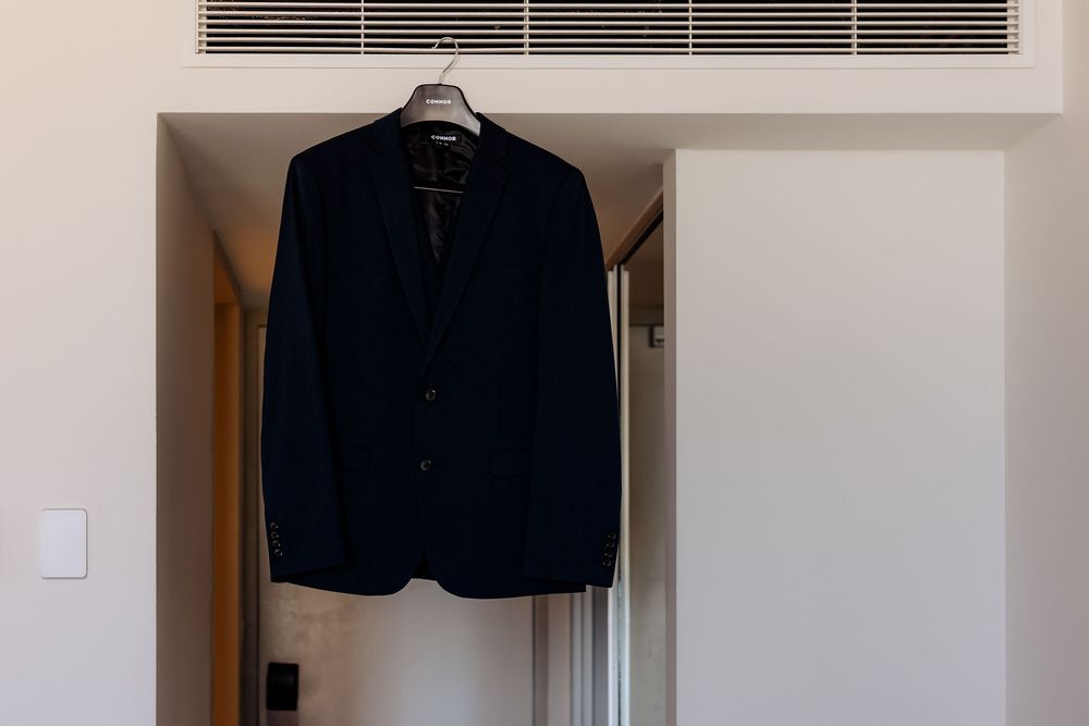 A black blazer jacket hangs on a hanger against a beige wall beneath an air conditioning vent.